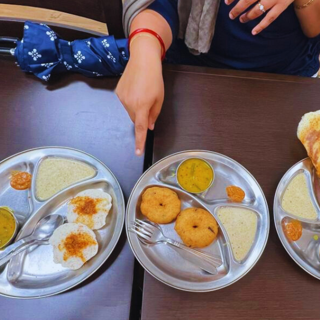 Finger pointing to two plates of South Indian food. One plate contains Steamed Idli and another plate contains Medu Vada. There's also coconut chutney and Sambhar on the side.